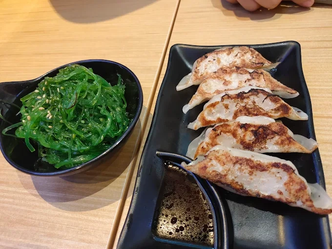a plate of five browned gyoza dumplings and a bowl of green seaweed salad sit on a wooden table. a small dish of dipping sauce accompanies the gyoza.