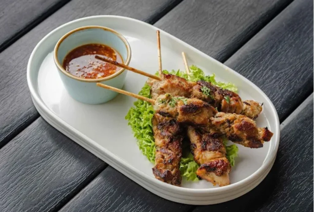 grilled chicken skewers on lettuce served with a small bowl of spicy red sauce on a white oval plate, placed on a dark wooden table.