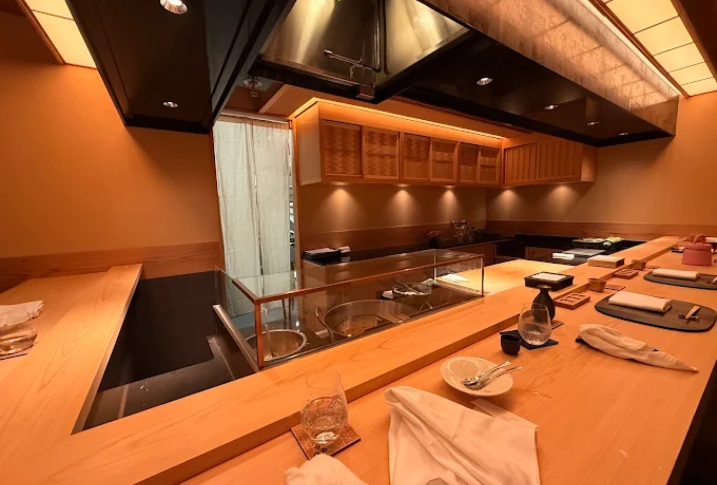 warmly lit sushi bar with a wooden counter and neatly arranged plates, cups, and menus. soft ambient lighting creates a cozy, inviting atmosphere.